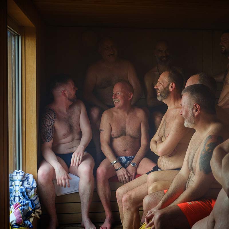 Scottish-Seaside-Saunas-Benefits-04 A group of men in swimwear sit closely together, talking and laughing inside a wooden sauna with a window on the left.